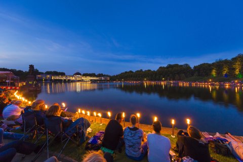Menschen sitzen mit Picknickdecken um den Hollersee und lauschen einem klassischen Konzert Menschen sitzen mit Picknickdecken um den Hollersee und lauschen einem klassischen Konzert